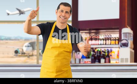 Hispanic barista wearing employee yellow apron success sign doing positive gesture with hand, thumbs up smiling and happy. Cheerful expression and win Stock Photo