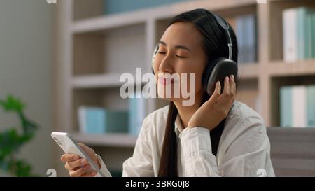 Happy smiling Asian Japanese businesswoman female Chinese Korean business woman girl student in wireless headphones listening music using mobile phone Stock Photo