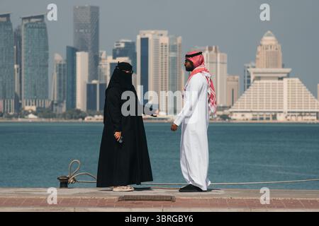 A couple stands by the water, enjoying the sunny weather. A man and a woman in traditional Arabic clothes walk along the embankment against the backdr Stock Photo