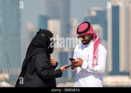 a man and a woman in traditional Arab clothes chat and laugh on the embankment against the backdrop of the sea and skyscrapers of Doha in Qatar. Febru Stock Photo