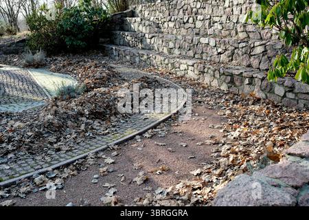A serene pathway covered in leaves winds alongside a textured stone wall. Stock Photo