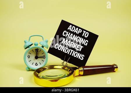 Adapt To Changing Market Conditions text message on sticky note with magnifying glass and alarm clock on yellow background Stock Photo