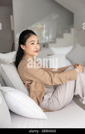 Thoughtful Asian woman dressed casually sits on cozy white couch Stock Photo