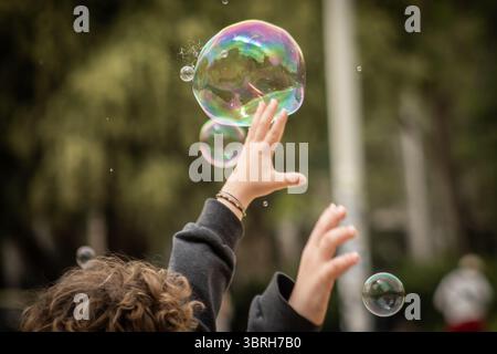 Kid enjoying a playful moment in nature, reaching out to touch iridescent soap bubbles floating in the air, creating a magical and carefree atmosphere Stock Photo