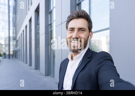A smiling businessman in a suit takes a selfie outside a modern building, wearing earbuds. Stock Photo