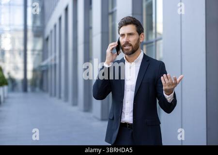 A businessman in a suit talks on his phone near a modern building. He gestures with his hand while speaking. Stock Photo