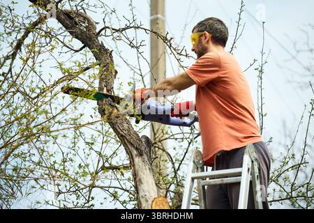 Young man cutting trees using electrical chainsaw in motion at his backyard. Gardening concept Stock Photo