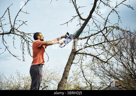 Young man cutting trees using electrical chainsaw in motion at his backyard. Gardening concept Stock Photo