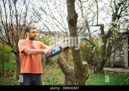 Young man cutting trees using electrical chainsaw in motion at his backyard. Gardening concept Stock Photo