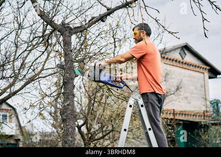 Young man cutting trees using electrical chainsaw in motion at his backyard. Gardening concept Stock Photo