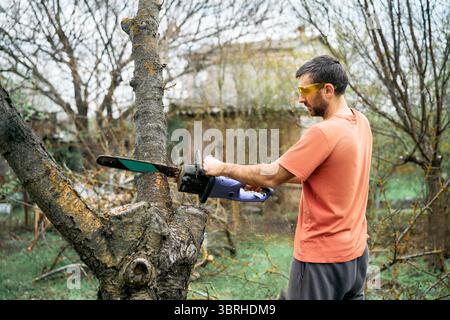 Young man cutting trees using electrical chainsaw in motion at his backyard. Gardening concept Stock Photo
