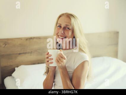 Portrait of happy smiling middle-aged woman enjoying hot tasty cup of coffee or tea in early morning in room at home Stock Photo