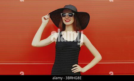 Portrait of beautiful young woman wearing a black summer hat and dress on red background Stock Photo
