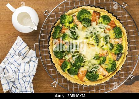 Top view of freshly baked delicious homemade broccoli and salmon quiche in black tart pan on metal cooling rack with white ceramic milk pot on wooden Stock Photo