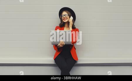Businesswoman working with laptop calling on phone wearing red blazer jacket, eyeglasses, round hat in the city on gray background, happy elegant woma Stock Photo