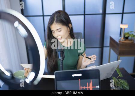 Happy young female making audio podcast in studio asian woman recording podcast on tablet with headphones and microscope for online streaming Stock Photo