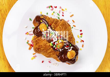 Croissant garnished with chocolate and rainbow sprinkles. Top view of delicious french croissant garnished with chocolate and rainbow sprinkles in whi Stock Photo