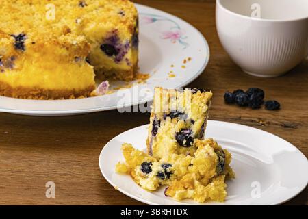 Piece of delicious homemade blueberry crumble cheesecake in white ceramic dish with the whole of them in ceramic plate and a coffee cup on wooden tabl Stock Photo