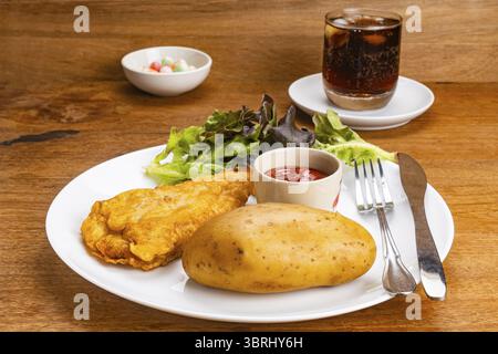 Closeup view battered fried fish, boiled potato, fresh green lettuce, a cup of ketchup, metal fork and table knife in white ceramic plate with glass o Stock Photo