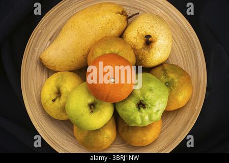 Top view pile of ripe fresh guava frui, red tomato, oranges, pear and apples in bamboo plate on black background Stock Photo