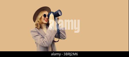 Portrait of stylish happy smiling woman photographer with digital camera taking picture wearing round hat, coat on studio background, blank copy space Stock Photo