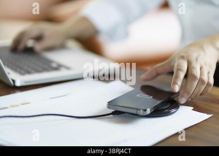 Charging mobile phone battery with wireless charging device in the table. Smartphone charging on a charging pad. Mobile phone near wireless charger Mo Stock Photo