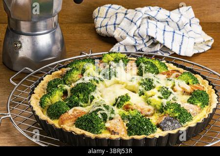 Freshly baked broccoli and salmon homemade quiche in black tart pan on metal cooling rack with moka pot coffee maker on wooden table Stock Photo