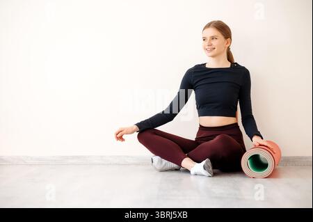 Attractive positive fit woman sitting on floor in lotus position and touching yoga mat with hand. Slim lady dressed in sport uniform looking aside and Stock Photo