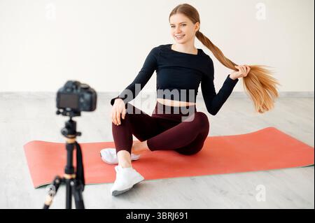 Athletic female blogger in sportswear shooting video content on camera and demonstrating online exercises. Charming lady with long ponytail sitting on Stock Photo