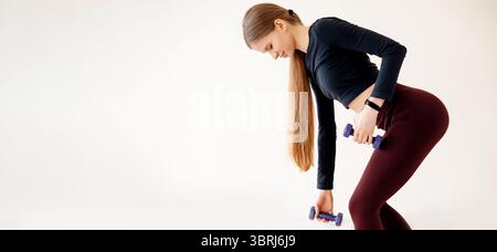 Slender female with perfect body shape doing regular workout with sport equipment indoors. Fit woman training with dumbbells and practicing romanian d Stock Photo