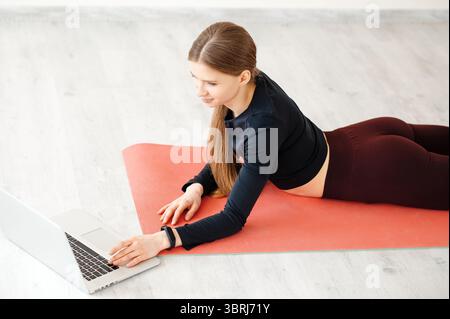 Closeup of young brunette woman in sportswear laying on fitness mat and using laptop, typing on keyboard, choosing online video for training from home Stock Photo