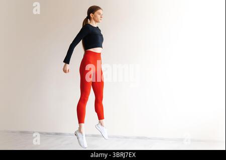 Active healthy woman doing cardio exercises and jumping in front of windows of white studio. Young energetic female dressed in tight sports clothes tr Stock Photo