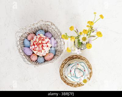 Sweet Easter cakes, colorful Easter eggs in the wooden baskets and yellow Ranunculus asiaticus in the glass vase on the white background. Top view Stock Photo