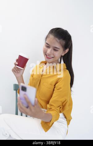 Asian woman holding phone and coffee cup with yellow shirt on white background Stock Photo