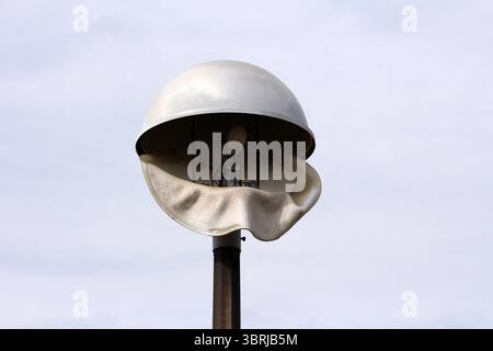 A street lamp features a white dome shade warped and melted, exposing bulbs and wiring, highlighting damage from heat or fire against a cloudy sky Stock Photo