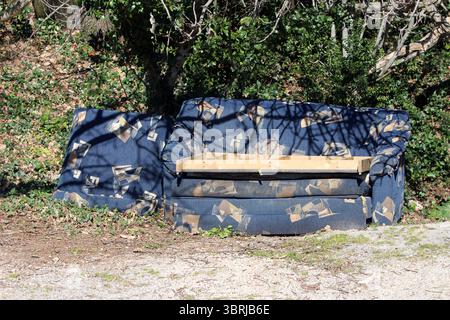 A worn, blue patterned sofa with exposed wooden frame and torn upholstery lies abandoned among grass and ivy, partially shaded by trees in a neglected Stock Photo