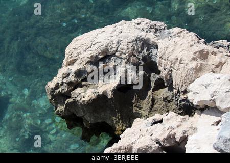A rugged limestone rock juts over transparent blue water, its weathered surface displaying sharp angles and natural crevices, evoking the raw beauty Stock Photo