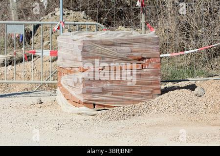 A large stack of red clay bricks is tightly wrapped in clear plastic film, resting on a construction site surrounded by gravel, fencing, and safety Stock Photo