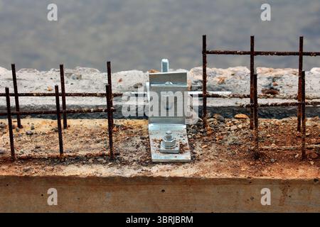 A galvanized steel anchor bracket is bolted to a concrete beam, surrounded by exposed and rusted rebar, highlighting early construction stages Stock Photo