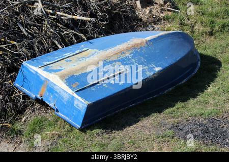 A faded blue rowboat with peeling paint and rust stains lies overturned on a grassy bank, showing clear signs of age and exposure near a pile of dry Stock Photo