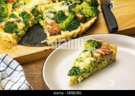 High angle view of freshly baked delicious homemade broccoli and salmon quiche on wooden cutting board and a slice in brown ceramic dish on wooden tab Stock Photo