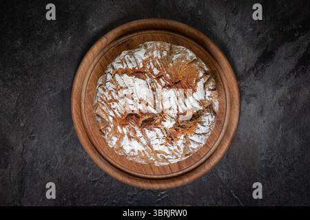 Round rustic loaf of bread baked in a Dutch oven, placed on circular wooden board, top-down view on dark slate background, home baking concept. Stock Photo