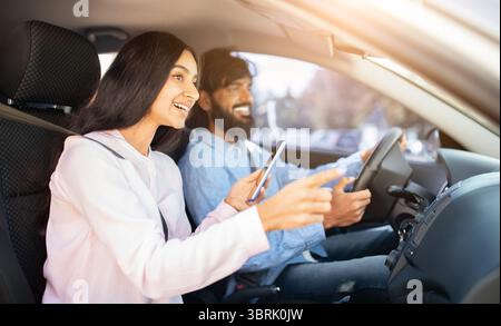 Cheerful Indian woman pointing out window with phone while man driving car Stock Photo