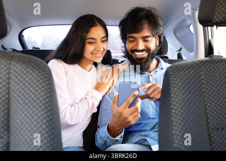 Young smiling indian couple sitting in car backseat, using smartphone together Stock Photo