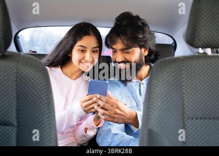 Smiling Indian couple sitting in backseat, happily browsing smartphone together Stock Photo