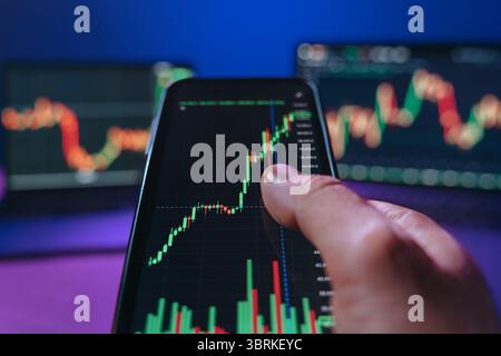 Close-up, trader holding his phone and analyzing stock and cryptocurrency movements using a trading app in front of his desk equipped with multiple monitors to track market trends. Stock Photo
