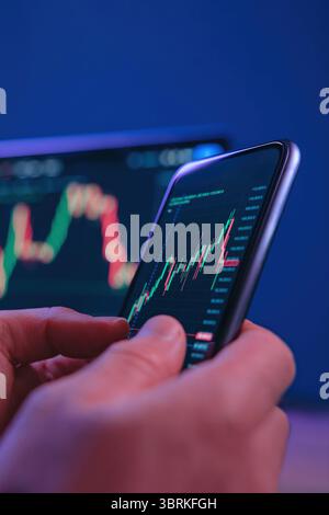 Close up, traders or investors hands using smartphone for real-time stock market monitoring with candlestick chart in a modern tech setup with neon light. Trading app with crypto graphs on screen Stock Photo