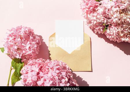 Blank white card in kraft envelope with pink hydrangea flowers on pastel background. Top view, flat lay, mockup. Stock Photo