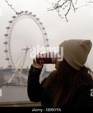 A woman in a beige beanie and black jacket sips from a red coffee cup with the London Eye in the background on a foggy day. Stock Photo