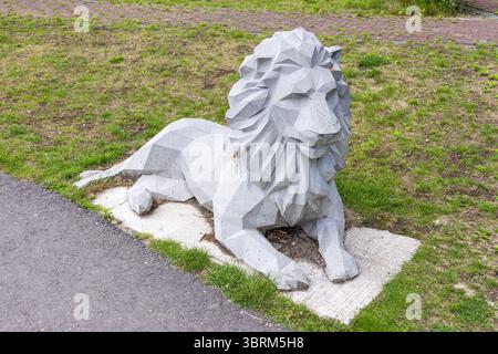 An angular, geometric concrete lion sculpture,  rests on a concrete slab amidst green grass and a paved path, showcasing modern public art. Stock Photo